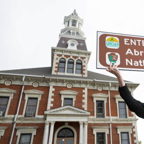 Person in Lincoln-era costume holding a "Entering Abraham Lincoln Natl Heritage Area" sign with historic building backdrop.