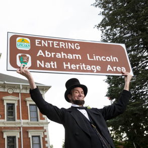 Person in Lincoln-era costume holding a "Entering Abraham Lincoln Natl Heritage Area" sign with historic building backdrop.