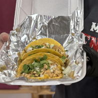 Three tacos in a takeout container, filled with chicken, cilantro, and onions in yellow corn tortillas, evoke comfort and craving.