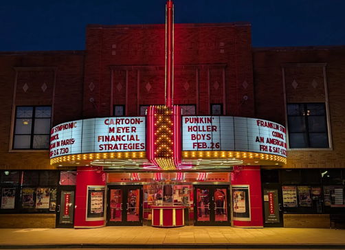 Vintage theater with red neon lights at twilight, marquee showcasing events, evoking nostalgia and excitement in a lively atmosphere.