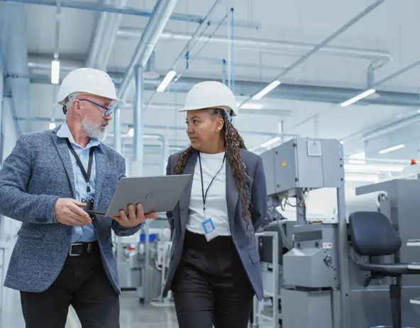 Two individuals in hard hats discuss over a laptop in a clean, modern industrial facility, showcasing teamwork and professionalism.
