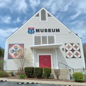 Museum with a triangular roof and white exterior, vibrant artworks, red door, surrounded by greenery under a blue sky.