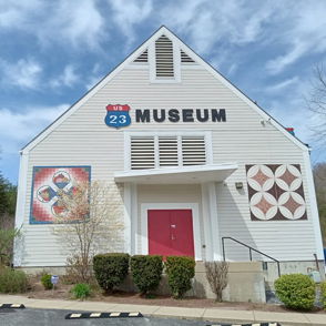 Museum with a triangular roof and white exterior, vibrant artworks, red door, surrounded by greenery under a blue sky.