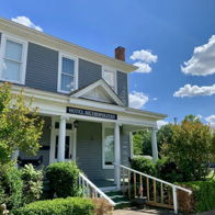 A charming gray house with white trim and a front porch, surrounded by greenery, under a sunny blue sky. Inviting and nostalgic.