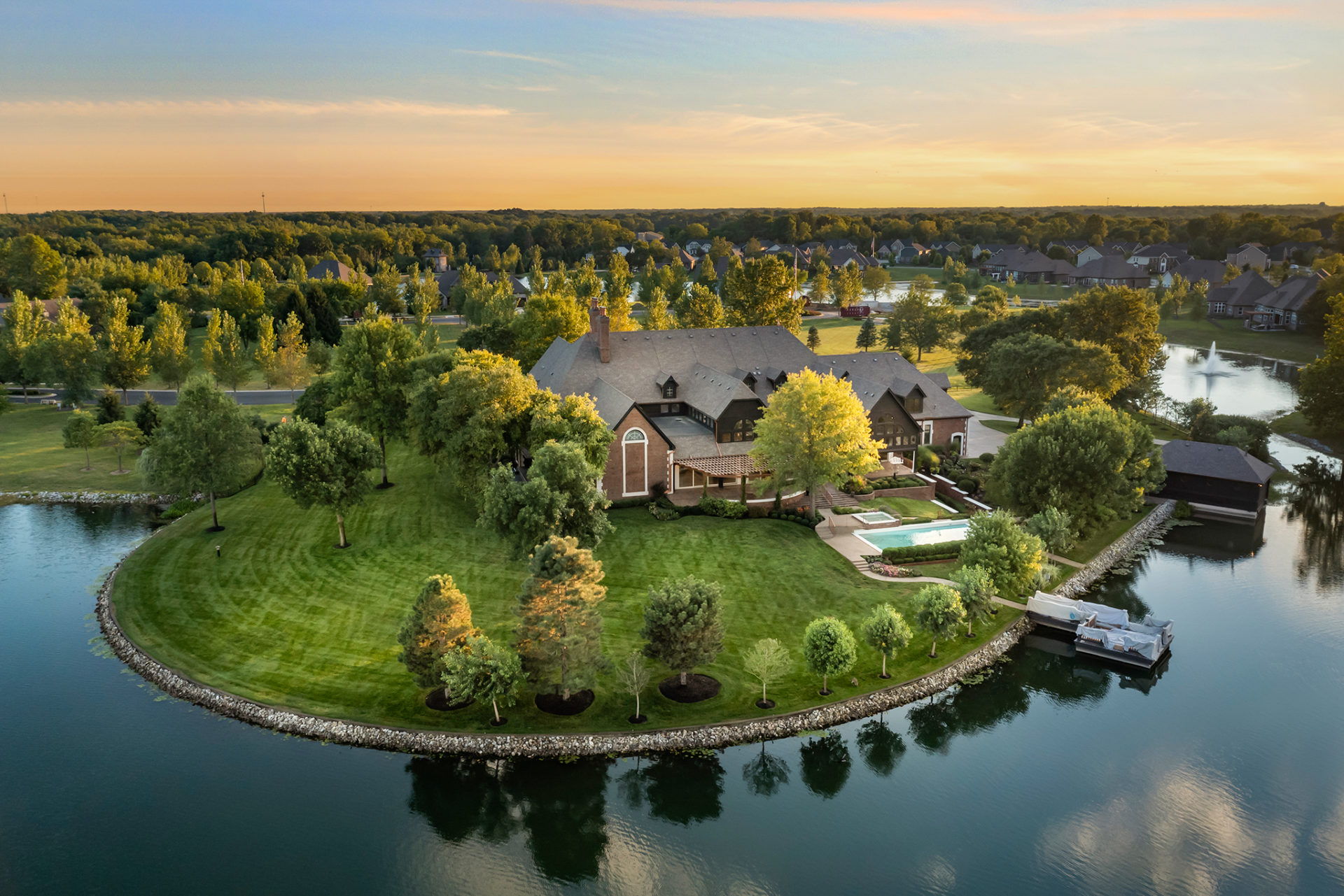 Aerial view of a serene house on a lush island, featuring a pool, surrounded by greenery and reflecting a warm sunset.