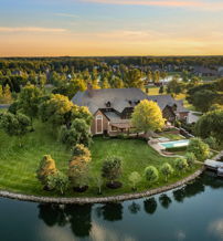 Aerial view of a serene house on a lush island, featuring a pool, surrounded by greenery and reflecting a warm sunset.