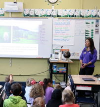 A teacher engages attentive young students in a bright classroom, presenting nature-related images on a projector screen.