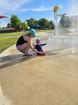 Joyful scene at a splash pad: a parent crouches by a fascinated child playing with water under a sunny sky.