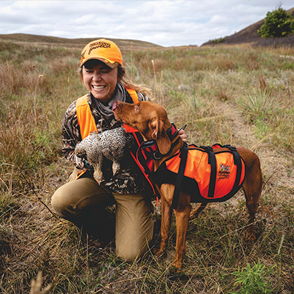 A joyful person in outdoor gear kneels with a dog and bird, highlighting a bond and love for nature and adventure.