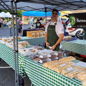 A friendly vendor showcases baked goods at a vibrant farmers' market, surrounded by colorful stalls and a lively atmosphere.