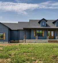 Modern blue house with stone accents, large windows, and a porch, set in a serene grassy area under a bright sky.