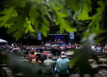Outdoor concert in a natural setting with performers and an audience on benches, evoking joy and community amidst greenery.