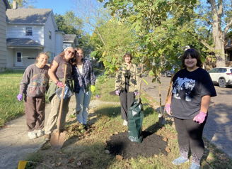 Five people joyfully plant a young tree together, embodying community spirit and hope for the environment on a sunny day.