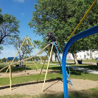 Playground with blue sky, colorful play structure, climbing frame, swing set, and a person adjusting equipment; joyful and playful scene.