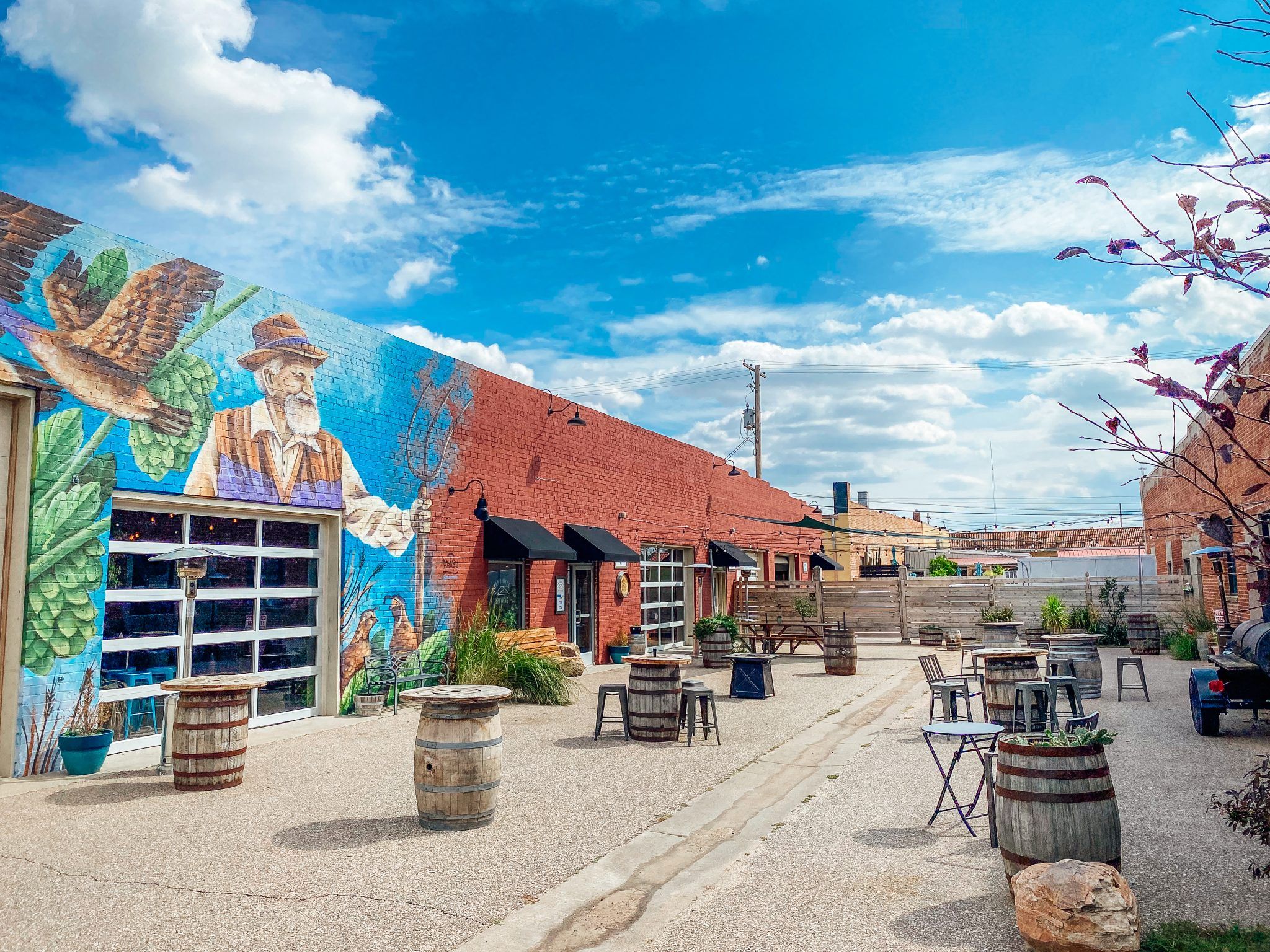 Vibrant mural and gathering space with barrels and tables under a blue sky, evoking joy, community, and relaxation.