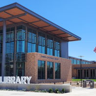 Modern library with glass windows, stone and wood design, "LIBRARY" sign, American flag, in a bright, sunny setting.