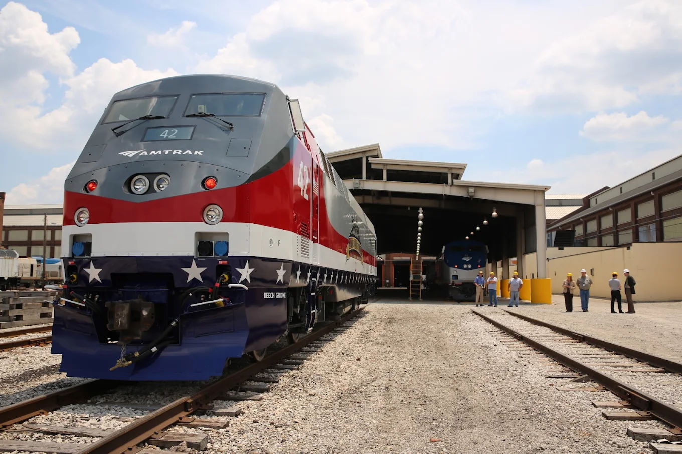 Modern train in red, white, and blue at tracks near service facility, with workers symbolizing teamwork and patriotic pride.