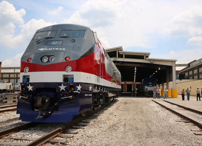 Modern train in red, white, and blue at tracks near service facility, with workers symbolizing teamwork and patriotic pride.