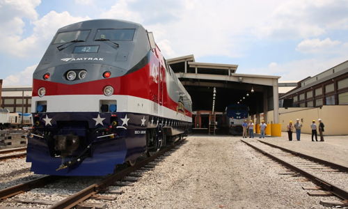 Modern train in red, white, and blue at tracks near service facility, with workers symbolizing teamwork and patriotic pride.