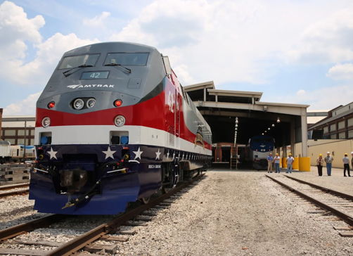 Modern train in red, white, and blue at tracks near service facility, with workers symbolizing teamwork and patriotic pride.