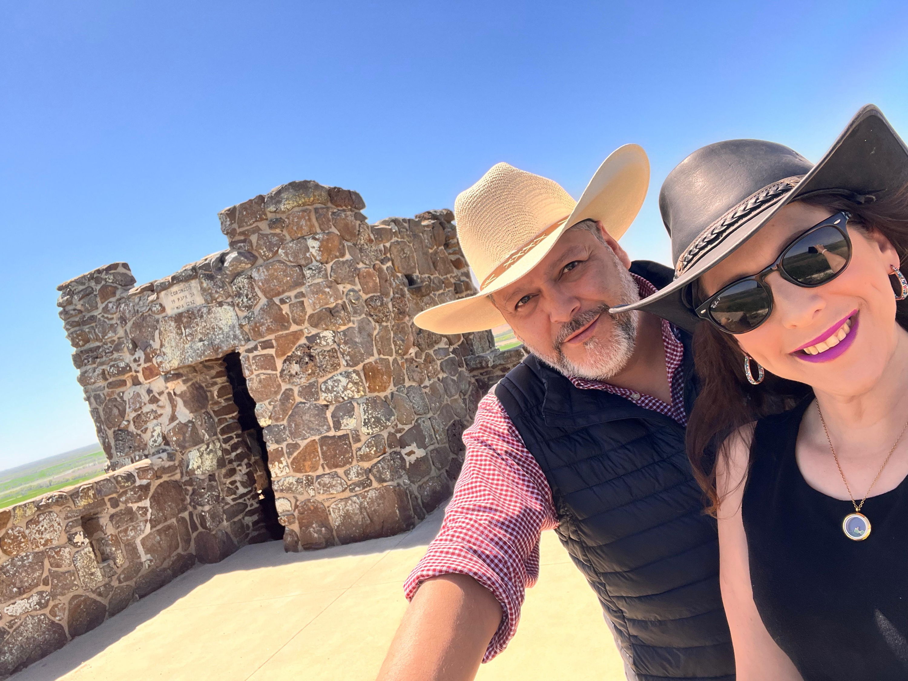 Two smiling individuals outdoors in front of a stone structure, radiating joy and adventure in a sunny setting.