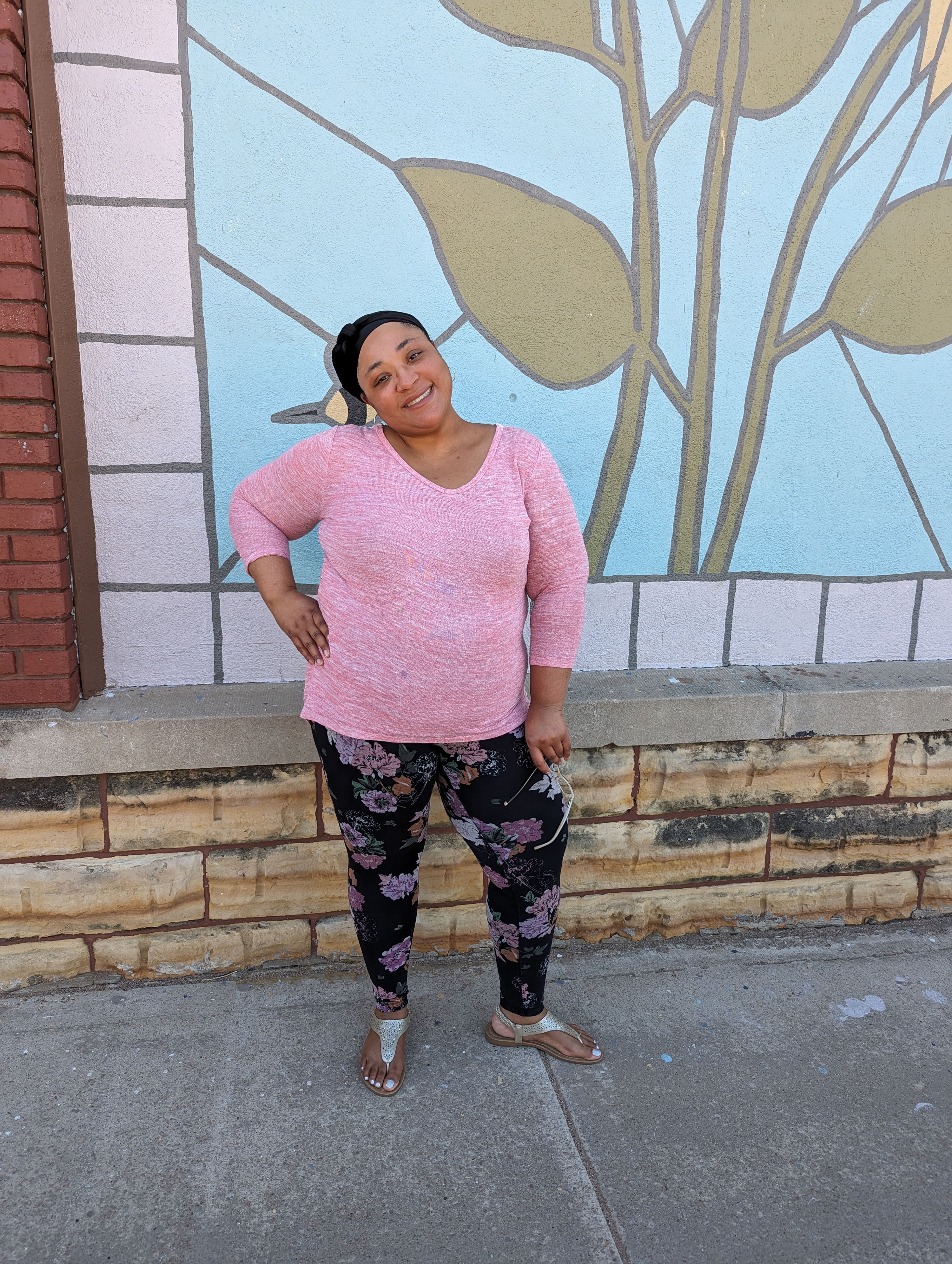 A woman in a pink sweater stands confidently by a colorful leaf mural, exuding happiness and a cheerful atmosphere.