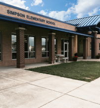 Elementary school with a brick facade, blue roof, large windows, grass, walkway, and potted plants, conveying warmth and community.
