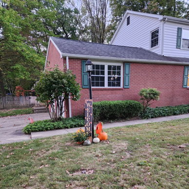 Cozy brick house with green shutters, pumpkins, and seasonal decor, evoking warmth and nostalgia for fall.