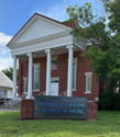 Charming brick building with white columns, gabled roof, and a friendly sign, set in a green, inviting environment.