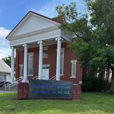 Charming brick building with white columns, gabled roof, and a friendly sign, set in a green, inviting environment.