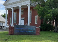 Charming brick building with white columns, gabled roof, and a friendly sign, set in a green, inviting environment.