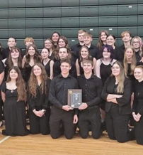 A joyful choir in black attire poses with a trophy in a gym, showcasing unity and pride after a successful performance.