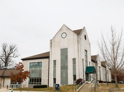Modern building with a pointed roof, clock, and neutral tones, surrounded by bare trees under an overcast sky, evoking calmness.