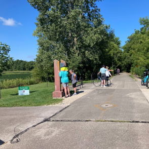 A peaceful park pathway with people biking, reading, and relaxing, surrounded by lush greenery under a sunny sky.