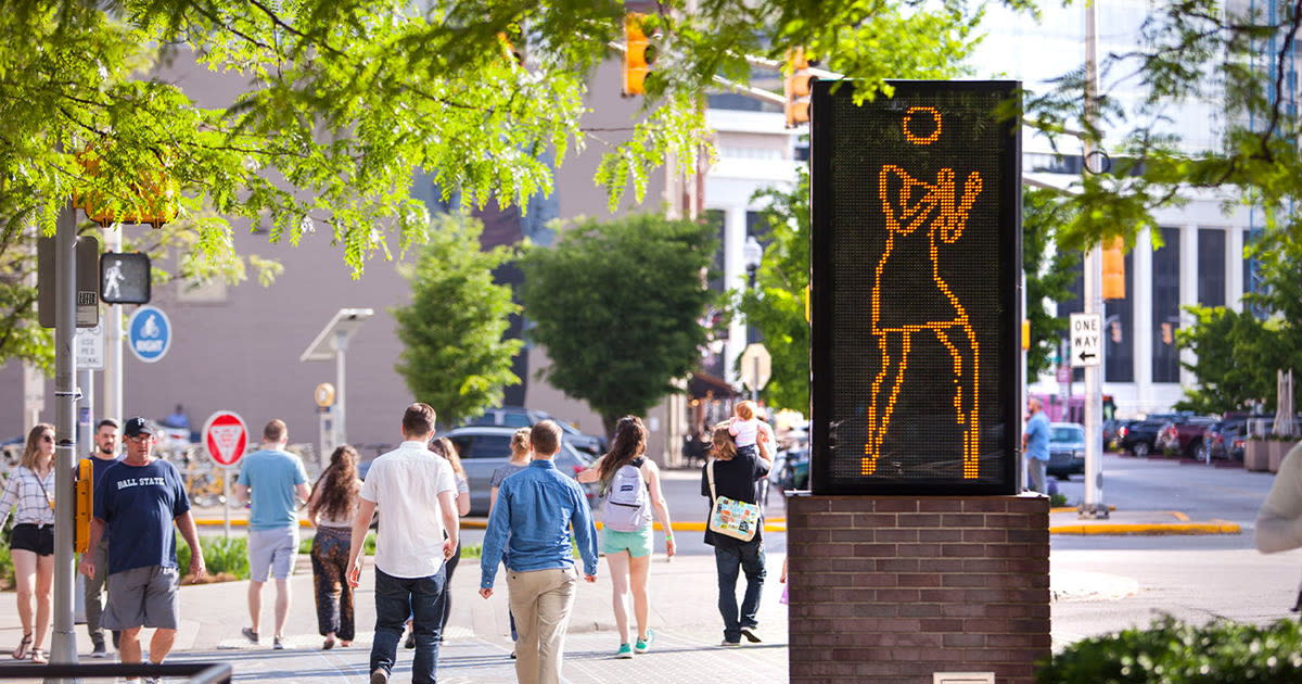 Vibrant street scene with people, a dynamic sculpture, greenery, and a cheerful atmosphere of community and celebration.