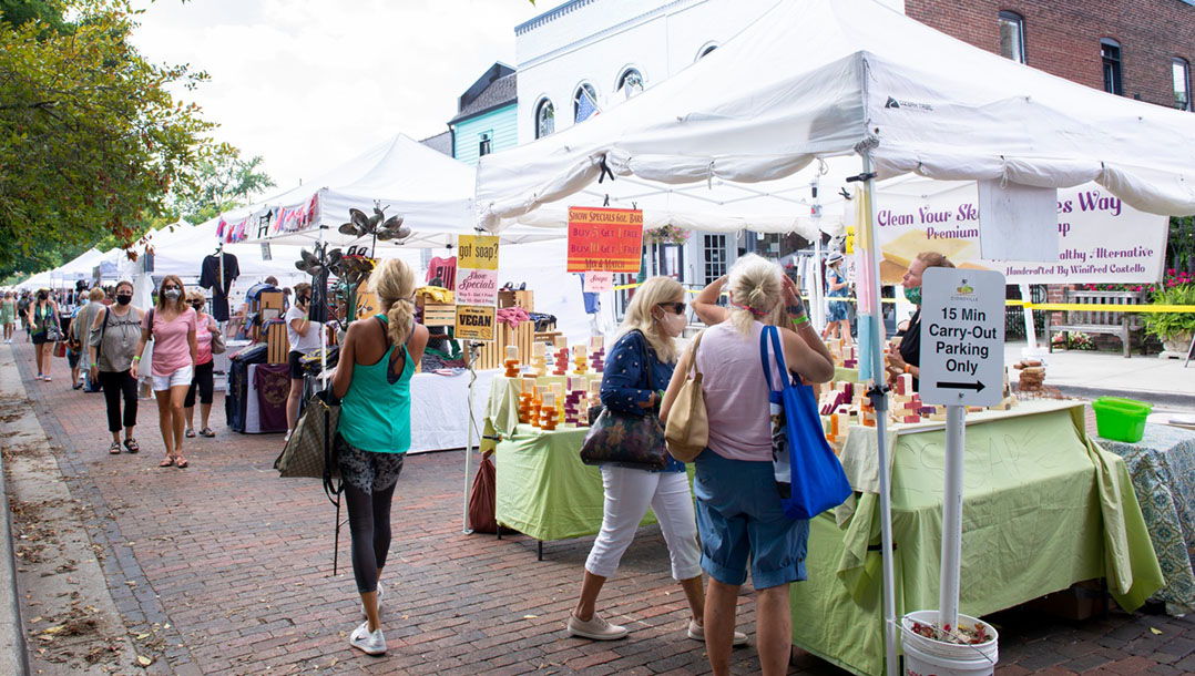 A lively outdoor market with white tents, colorful signs, and people browsing, evoking community joy and excitement.
