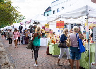 A lively outdoor market with white tents, colorful signs, and people browsing, evoking community joy and excitement.