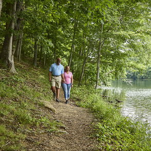 A couple strolls along a serene trail by water, surrounded by lush greenery, evoking calmness and companionship.