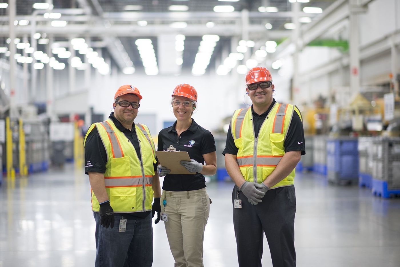 Three individuals in safety gear smile in a bright industrial space, conveying teamwork and a supportive work atmosphere.