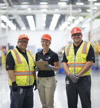 Three individuals in safety gear smile in a bright industrial space, conveying teamwork and a supportive work atmosphere.