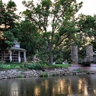 Serene outdoor scene with tranquil water, lush greenery, a cozy building, and a wooden bridge, evoking peace and relaxation.
