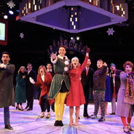 Festive theater scene with colorful costumes and joyful ensemble captures holiday spirit and community celebration.
