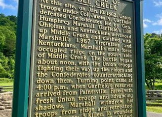 Historical marker for the Battle of Middle Creek, 1862, set against a serene backdrop of blue sky and green trees.