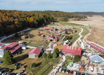 Aerial view of a festive autumn gathering with red-roofed buildings, lush trees, and a joyful crowd.