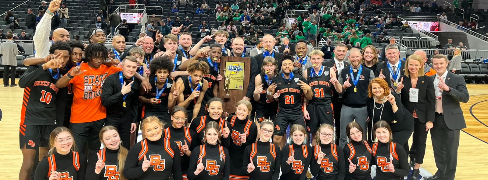 Basketball team and staff celebrate victory on court, holding a trophy, smiling, and showing camaraderie and joy.