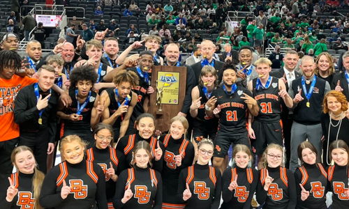 Basketball team and staff celebrate victory on court, holding a trophy, smiling, and showing camaraderie and joy.