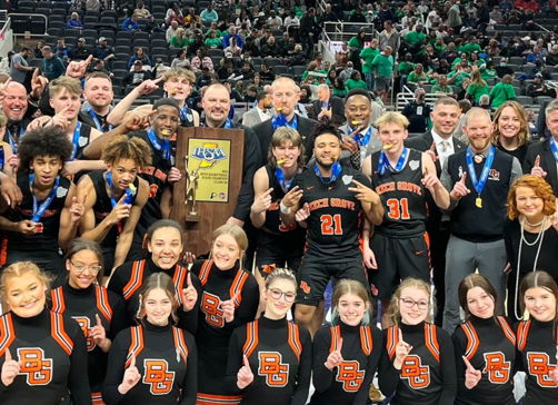Basketball team and staff celebrate victory on court, holding a trophy, smiling, and showing camaraderie and joy.