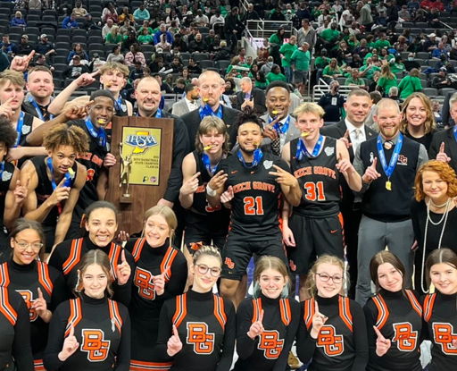 Basketball team and staff celebrate victory on court, holding a trophy, smiling, and showing camaraderie and joy.