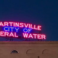 Neon sign "MARTINSVILLE CITY OF MINERAL WATER" glows against twilight, evoking nostalgia and community spirit.