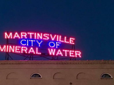 Neon sign "MARTINSVILLE CITY OF MINERAL WATER" glows against twilight, evoking nostalgia and community spirit.